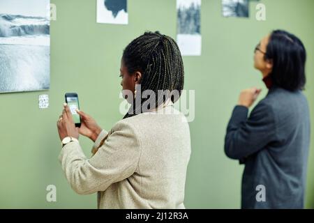 Junge afroamerikanische Frau besucht Ausstellung in der Galerie für moderne Kunst Scannen QR-Code mit dem Smartphone, um mehr Informationen über Foto an der Wand zu finden Stockfoto