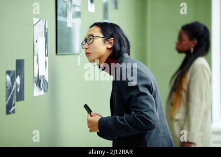 Porträt eines hübschen jungen asiatischen Mannes mit Brille, der eine Ausstellung in einer Kunstgalerie besucht, die sich auf ein Schwarzweiß-Foto an der Wand konzentriert Stockfoto