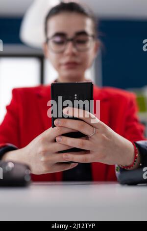 Nahaufnahme einer Geschäftsfrau, die das Smartphone über den Schreibtisch hält. Hände des Unternehmers mit Brille mit Smartphone in Startup-Büro. Inhaber eines Kleinunternehmens in roter Jacke, der Inhalte auf mobilen Geräten durchsucht. Stockfoto