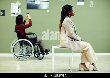 Nicht erkennbarer junger Mann mit Behinderung und schwarze Frau, die die Ausstellung zeitgenössischer Fotografie in der Kunstgalerie besucht Stockfoto