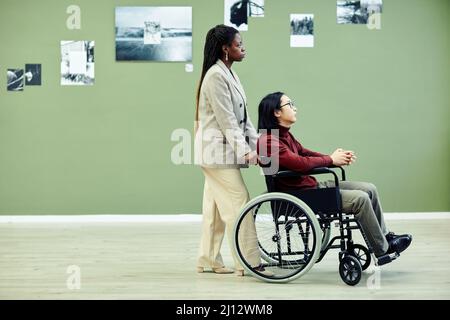 Stilvolle junge afroamerikanische Frau und ihr asiatischer Freund mit Behinderung im Rollstuhl besuchen zeitgenössische Fotoausstellung Stockfoto