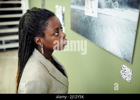 Mittelgroße Nahaufnahme einer nachdenklichen jungen schwarzen Frau mit Afro-Flechtfrisur, die eine zeitgenössische Ausstellung besucht und Fotos an der Wand ansieht Stockfoto