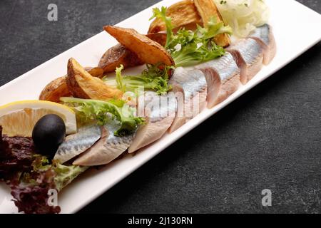 Eingelegter Hering mit gebackenen Kartoffeln, Zwiebeln und Salat auf dunklem Grund Stockfoto