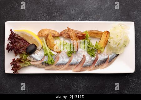 Eingelegter Hering mit gebackenen Kartoffeln, Zwiebeln und Salat auf dunklem Grund Stockfoto