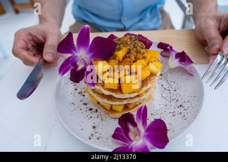 Leckere Pfannkuchen mit tropischen Früchten auf einem Teller auf einem Tisch in einem Café, Nahaufnahme. Gesundes Frühstück. Leckere Pfannkuchen mit Mango, Drachenfrucht und Pass Stockfoto