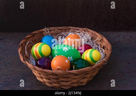 Geflochtener Korb mit bunt bemalten ostereiern auf einem Nest aus Holzwolle, isoliert von dunklem Hintergrund Stockfoto