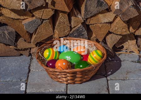 Geflochtener Korb mit bunten ostereiern in einem Nest aus Holzwolle, Blockstapel im Hintergrund, aus der Nähe und isoliert Stockfoto