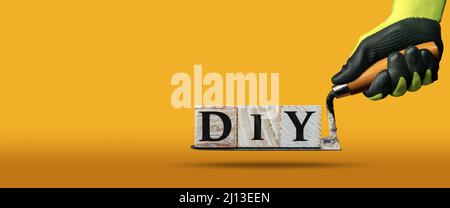 Arbeiter mit Arbeitsschutzhandschuhen, die eine schmutzige Kelle und den DIY-Text (do it yourself) halten, aus Holzblöcken. Gelber, orangefarbener Hintergrund. Stockfoto