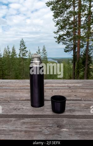 Dunkelblaue Thermoskanne aus Stahl und Becher aus Stahl auf einem Holztisch mit Naturhintergrund in den Wäldern Finnlands Stockfoto