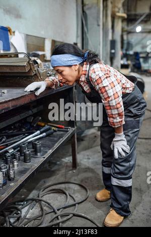 Junge Technikerin in Arbeitskleidung, die sich vor der Werkbank biegt, während sie Handwerkzeuge für die Reparatur von Industriemaschinen auswählt Stockfoto