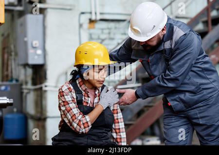 Vorarbeiter in Hardhut und Arbeitskleidung, die neben einer Ingenieurin stehen, wobei sie ihre Schulter mit der Maschine berührte Stockfoto