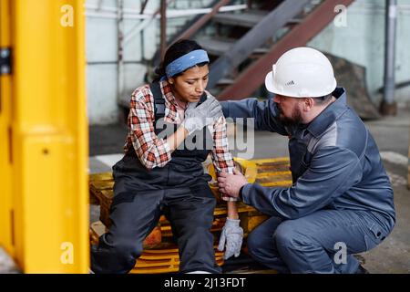 Reifer Vorarbeiter in Arbeitskleidung, der einer weiblichen Untergebenen erste Hilfe geleistet hat, mit Schmerzen in der Schulter nach einem Unfall bei der Arbeit im Lager Stockfoto