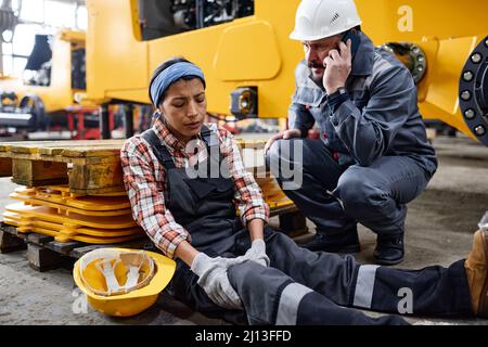 Junge Ingenieurin mit schmerzenden Knien, die von einem ängstlichen männlichen Arbeiter in Schutzhelm und Uniform auf dem Boden sitzen und den Krankenwagen rufen Stockfoto