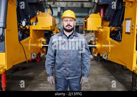 Selbstbewusster reifer männlicher Baumeister in Schutzhelm und Arbeitskleidung, der im Lager oder in der Werkstatt gegen riesige Industriemaschinen steht Stockfoto