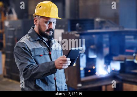 Selbstbewusster männlicher Ingenieur oder Baumeister in Arbeitskleidung mit digitalem Tablet zur Überprüfung von Daten über Waren, die von Online-Kunden bestellt wurden Stockfoto