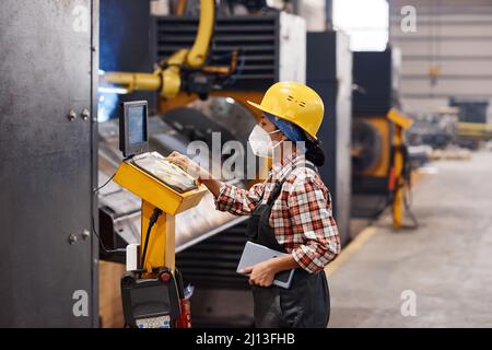 Seitenansicht einer jungen Ingenieurin in Atemschutzmaske, Hardhut und Overalls, die auf ein Dokument mit technischen Informationen in der Fabrik zeigt Stockfoto