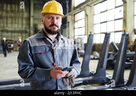 Seriöse reife Arbeiter oder Techniker mit Smartphone in Hardhat und Boilersuit stehen in großen Distributionslager Stockfoto