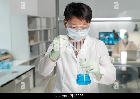 Junge asiatische Chemikerin, die bei der Durchführung wissenschaftlicher Experimente im Labor einen Tropfen der chemischen Substanz in die blaue Flüssigkeit einfügte Stockfoto