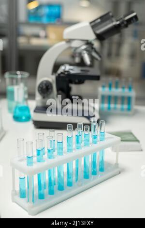 Die Gruppe der Kolben mit den Proben der blauen Flüssigkeit, die auf dem Arbeitsplatz des Wissenschaftlers gegen das Mikroskop und andere Glasanlagen stehen Stockfoto