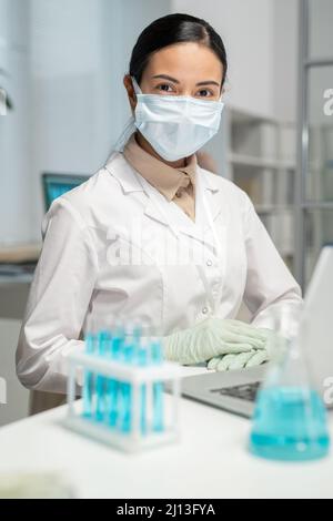 Junge Wissenschaftlerin in Whitecoat, Handschuhe und Schutzmaske sitzen am Schreibtisch vor dem Laptop und Set von Flaschen mit blauer Flüssigkeit Stockfoto