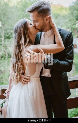 Braut und Bräutigam küssen, während sie auf dem Balkon eines Holzhauses im Wald stehen. Nahaufnahme im Hochformat. Herrliche Natur im Sommer. Stockfoto