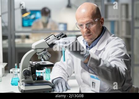 Moderner reifer männlicher Arzt mit Handschuhen, weißem Schaumstoff und Schutzbrille, der blaue Flüssigkeit im Kolben betrachtet, während er im Labor sitzt Stockfoto