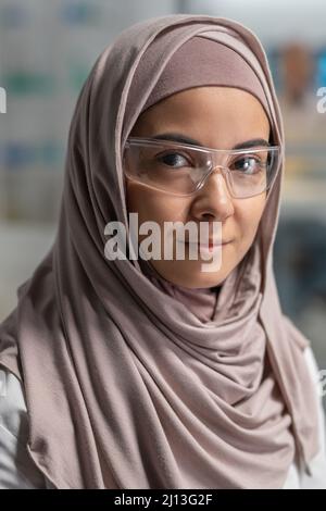 Junge erfolgreiche muslimische Forscherin in Hijab und Schutzbrille, die vor der Kamera steht und Sie anschaut Stockfoto