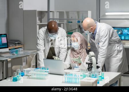 Junge muslimische Forscherin in Hijab, Whitecoat, Schutzhandschuhen, Maske und Brillen, die Präsentation vor männlichen Kollegen macht Stockfoto