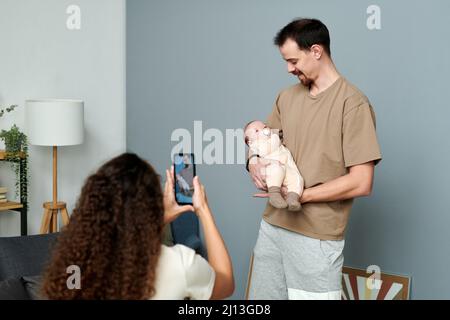 Glücklicher junger Mann, der auf den Händen einen kleinen Sohn ansieht, während er vor seiner Frau steht und sie auf dem Smartphone fotografiert Stockfoto