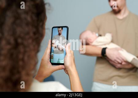 Vater hält den kleinen Sohn auf dem Bildschirm des Smartphones, das von einer jungen Frau gehalten wird, die ihren Mann und ihr neugeborenes Kind fotografiert Stockfoto