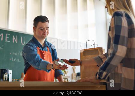 Fröhlicher junger Down-Syndrom-Kellner, der kontaktlose Smartphone-Zahlung vom Kunden im Take-Away-Restaurant nimmt. Stockfoto