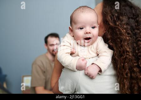 Glücklicher entzückender Baby Junge in weißem Strampelanzug auf den Händen seiner Mutter, die vor ihrem Mann zu Hause steht Stockfoto