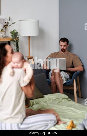 Seriös junger Mann mit Laptop-Networking, während er im Sessel vor seiner Frau mit ihrem entzückenden kleinen Sohn sitzt Stockfoto