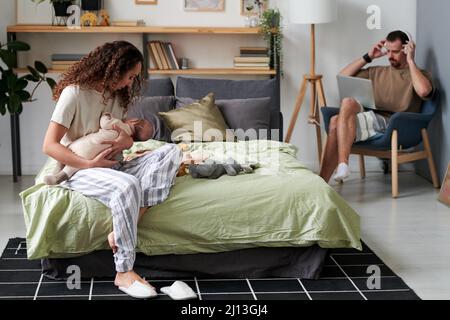 Junge Frau, die ein neugeborenes Baby schlafen lässt, während sie auf dem Doppelbett gegen ihren Mann sitzt und im Sessel ein Laptop-Netzwerk hat Stockfoto