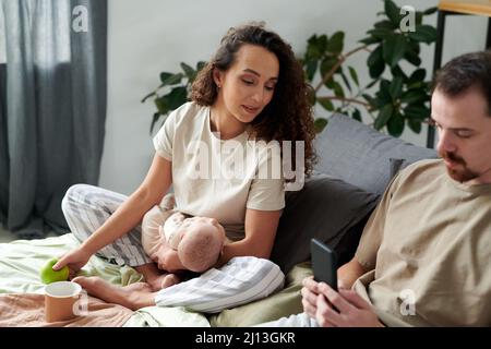 Junge Frau mit einem schlafenden kleinen Sohn auf den Händen, die im Bett frühstückte und auf den Bildschirm des Smartphones schaute, das von ihrem Mann gehalten wurde Stockfoto