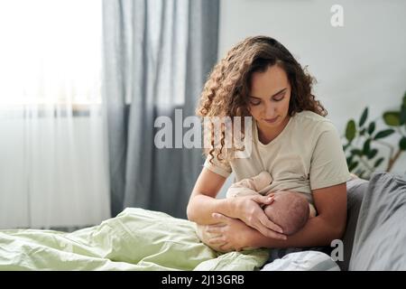 Junge, vorsichtige Mutter im Schlafanzug sitzt unter der Decke auf dem Bett und stillt ihr niedliches Baby, während sie ihn an den Händen hält Stockfoto