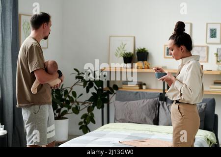 Ein junger Mann hielt einen schlafenden Sohn an den Händen, während seine vielbeschäftigte Frau mit einer Schüssel Essen im Schlafzimmer gegen ein Doppelbett frühstückte Stockfoto
