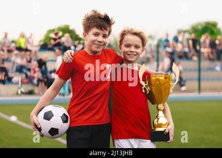 Glückliche Jungen Freunde Teamkollegen in der Fußballmannschaft gewinnen goldene Trophäe. Schulkinder in roten Fußballuniformen lächeln und halten den Ball und die goldene Tasse Stockfoto