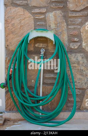 Grüner langer Gartenschlauch. Gewickelter Gartenschlauch, der am Wasserhahn an der Steinwand hängt. Vorderansicht, vertikal und Nahaufnahme. Stockfoto