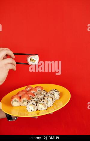 Nahaufnahme beschnittenes Foto von männlichem Halt in den Händen Makizushi Sushi-Rolle serviert auf dem Teller traditionelle japanische Küche isoliert auf rotem Hintergrund im Studio. Kopieren Stockfoto