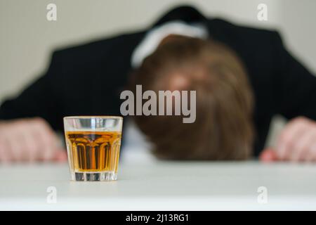 Betrunkener gestresster Geschäftsmann, der am Schreibtisch schläft. Alkoholabhängigkeit. Im Vordergrund ein Schuss Whisky. Stockfoto