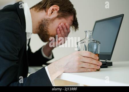 Müdigkeit junger Geschäftsmann sitzt am Arbeitsplatz und hält Whiskey-Flasche, Alkohol trinken, Konkurs-Konzept. Stockfoto