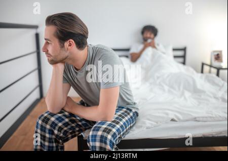 Gelaunter Kerl saß im Schlafzimmer weg von seinem Partner Stockfoto