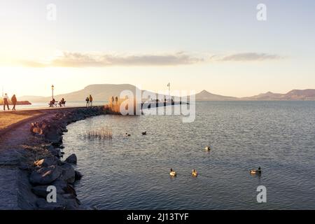 Touristen zu Fuß auf Fonyód Pier am Plattensee Winter Frühling Zeit in den Sonnenuntergang Badacsony Hügel Hintergrund. Stockfoto