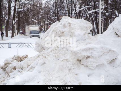 Eine hohe, große Schneeverwehung an der Straße vor dem Hintergrund einer Stadtstraße. Auf der Straße liegt schmutziger Schnee in hohen Haufen. Urbane Winterlandschaft. Wolkiger Wintertag, weiches Licht. Stockfoto