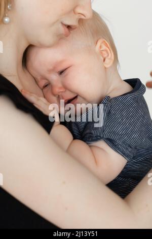 Gesicht des weinenden Kindes. Mutter fürsorglich, das keulende, depressive Baby in den Armen haltend. Temperatur, Krankheit, Kinderkolik. Nahaufnahme Stockfoto