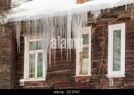 Mehrere transparente Eiszapfen hängen am Rand des Dachs. Vor dem Hintergrund der Holzwand des alten Hauses. Große Kaskaden, sogar schöne Reihen. Wolkiger Wintertag, weiches Licht. Stockfoto