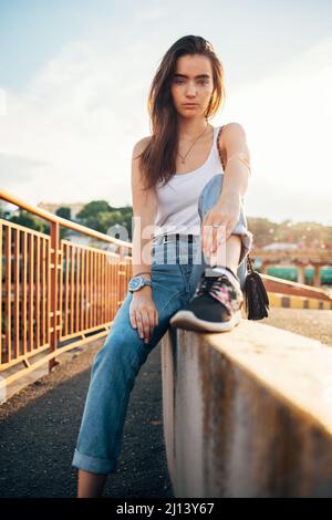 Portrait Mädchen mit langen dunklen Haaren tragen Jeans und weißen Top sitzen an der Stadtbrücke, sonnigen Sommertag. Stockfoto