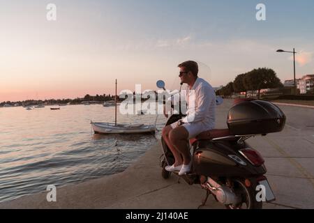 Ein moderner Geschäftsmann in legerer Kleidung sitzt auf einem Motorroller am Meer und genießt den Sonnenaufgang. Selektiver Fokus Stockfoto