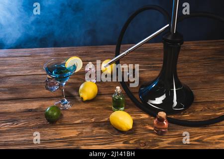 Shisha in der Nähe von Zitronen und Glas Cocktail auf Holzoberfläche auf schwarzem Hintergrund mit blauem Rauch Stockfoto